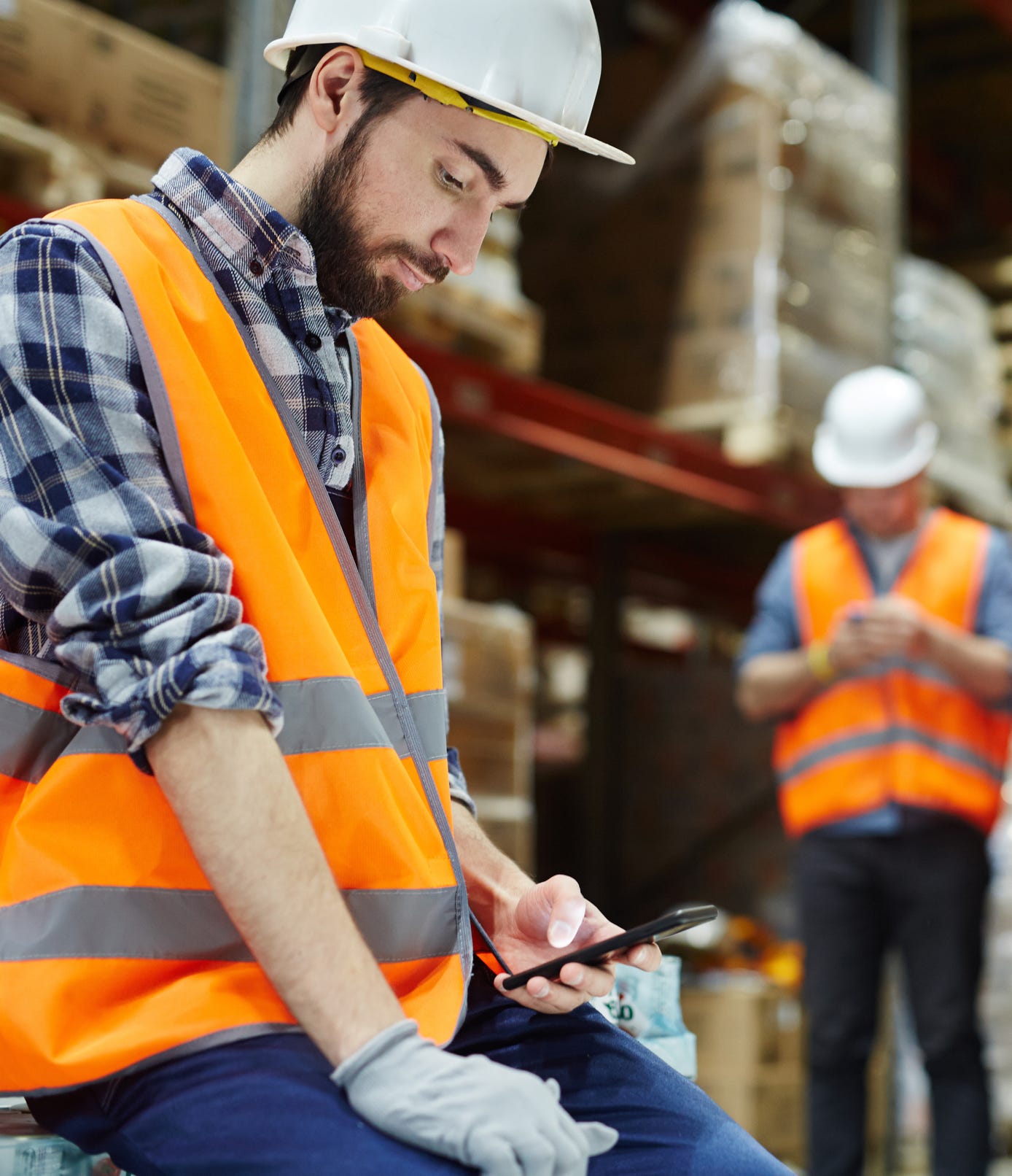 Warehouse worker in a hard hat and safety vest using his phone on the clock